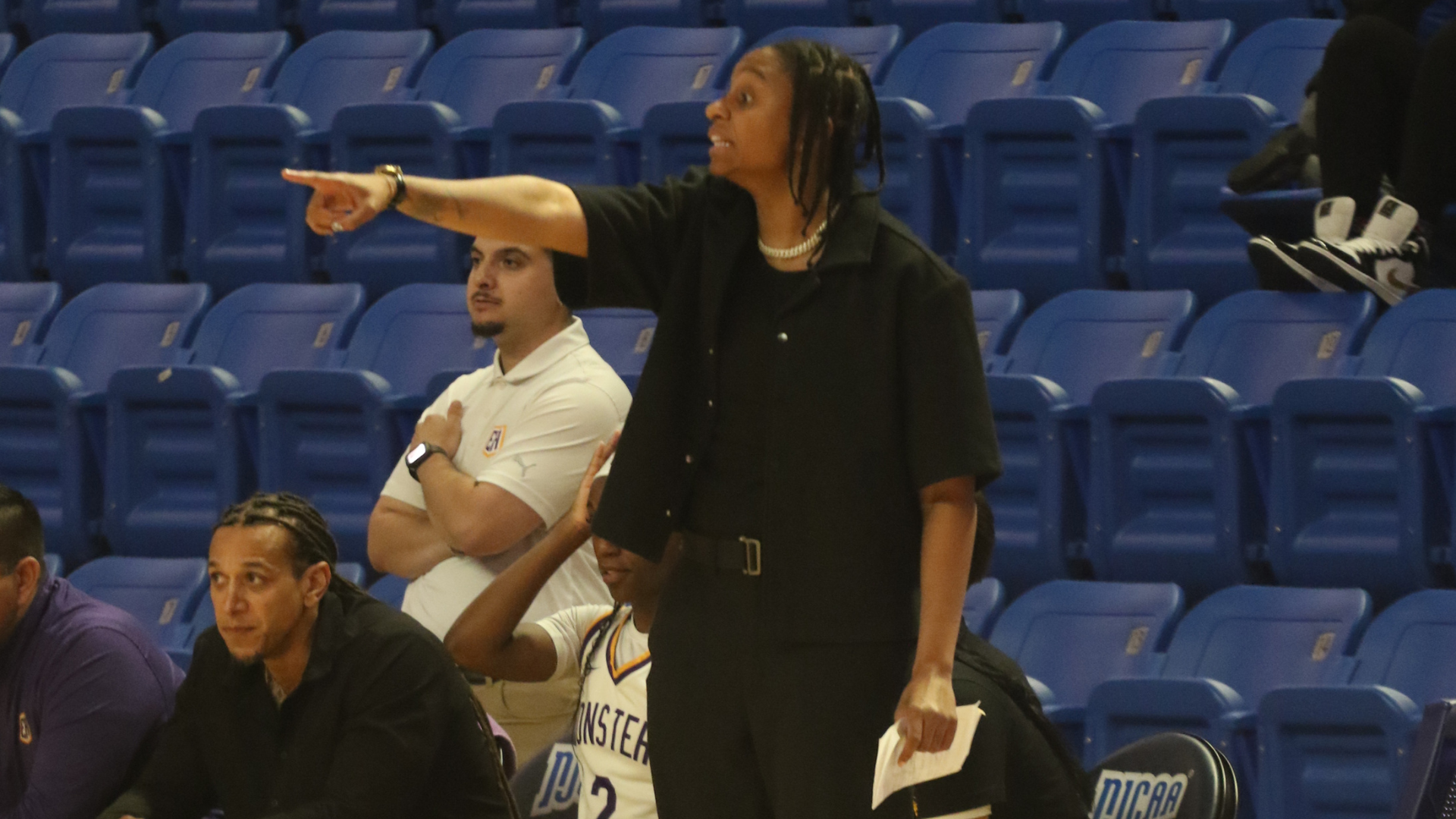 Eastern Arizona College Head Coach Angelica de Paulo coaching on the sidelines during a game with assistant coaches in the background. 